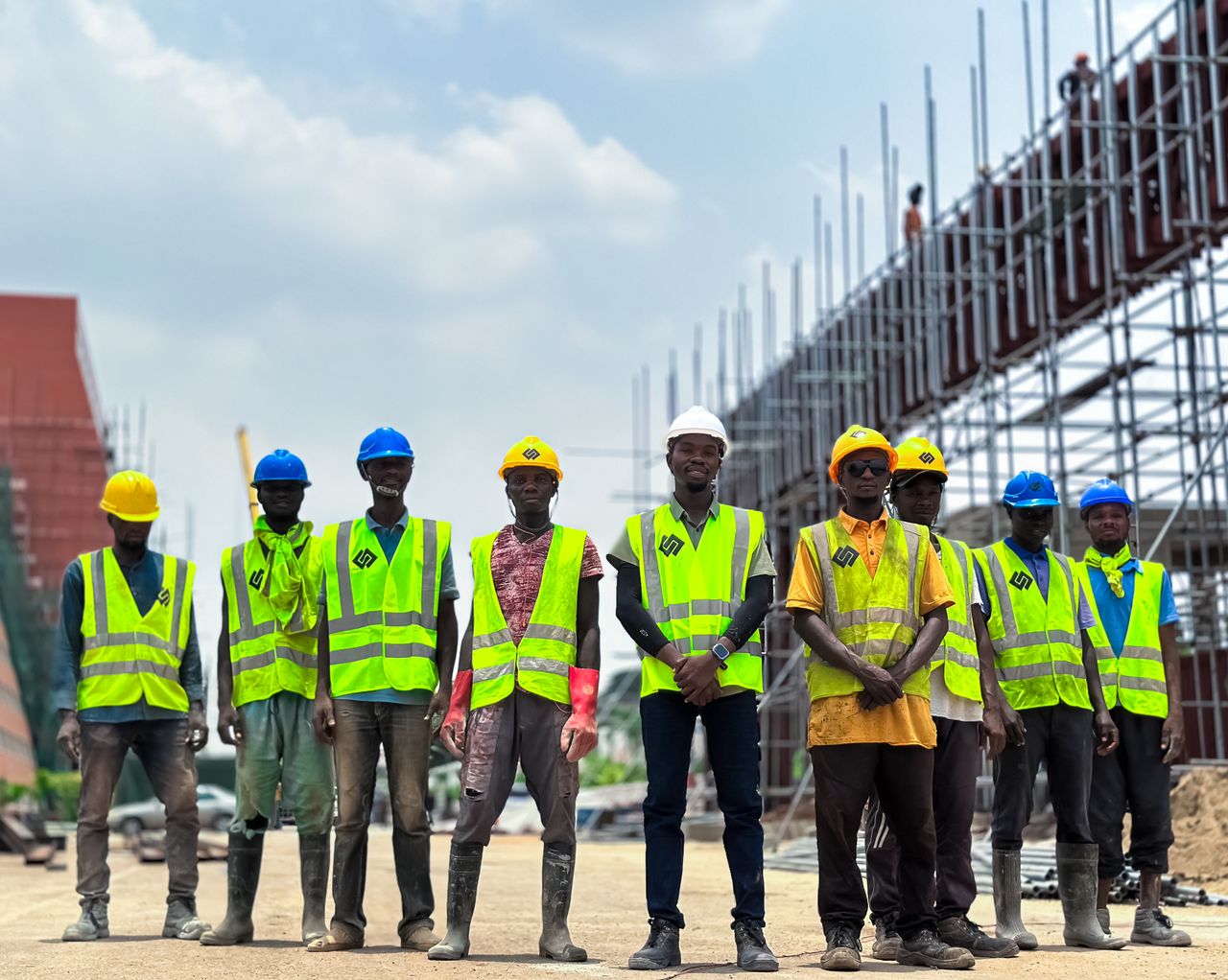 Group of workers in safety gear at a construction site. Visible scaffolding and clear sky.